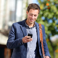 A person smiles while looking at a phone, carrying a bag, outdoors with trees and blurred buildings in the background.